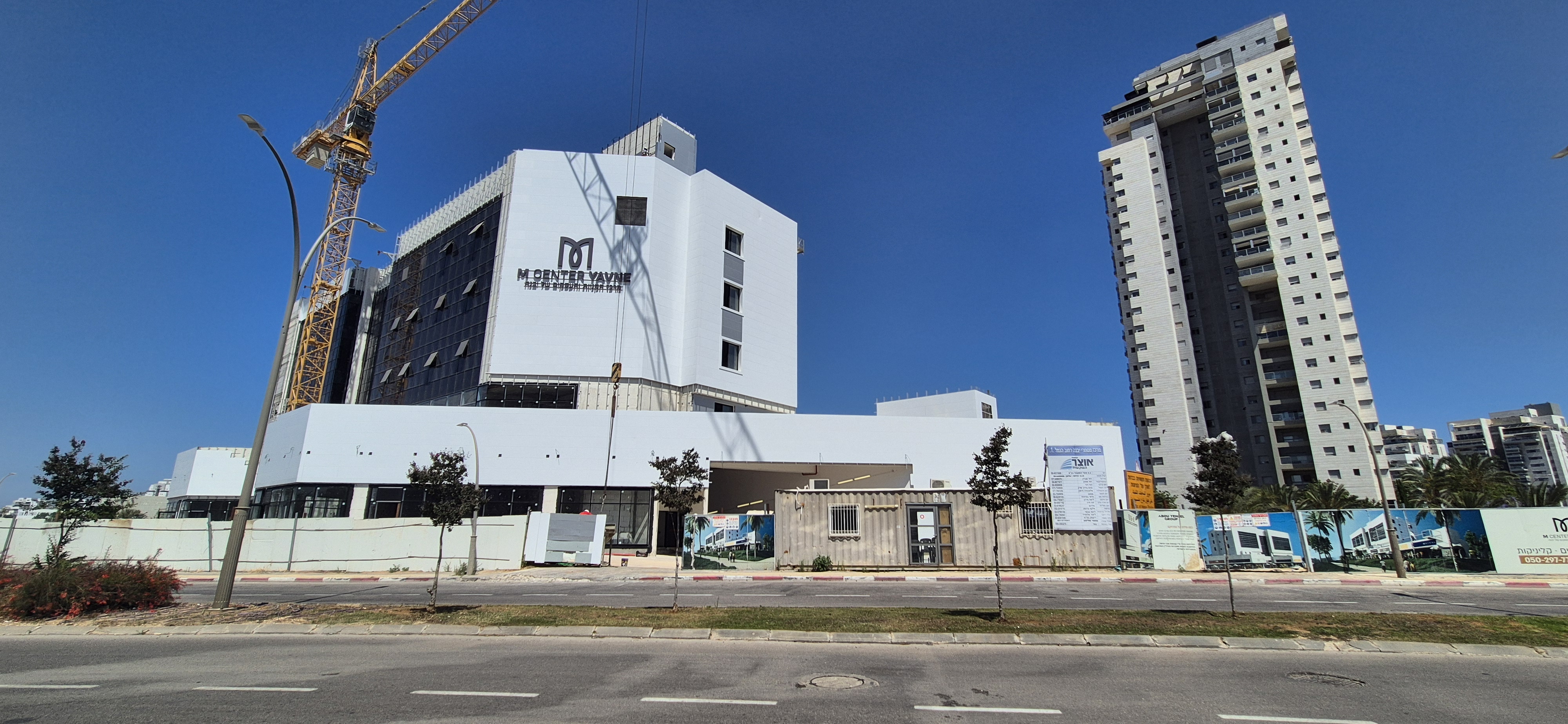 חזית M CENTER יבנה משדרות הציונות, עם לוגו המתחם ומגדל מגורים סמוך